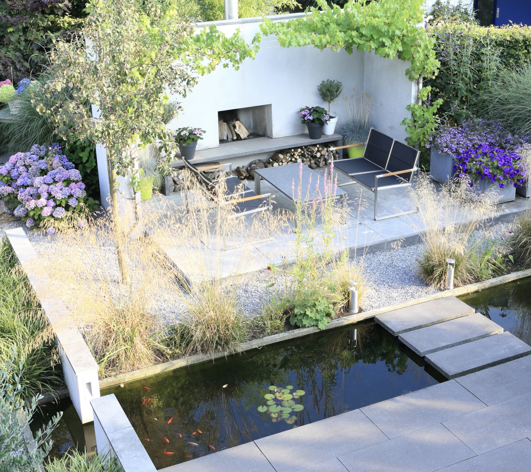 Overzicht van moderne tuin met witte buitenhaard, vijver met stapstenen en loungeterras