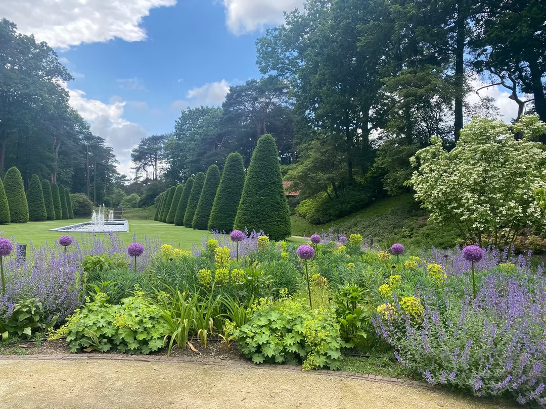 Formele landgoedtuin met kleurrijke vaste plantenborder, paarse Allium, waterpartij en kegelvormige taxusbomen