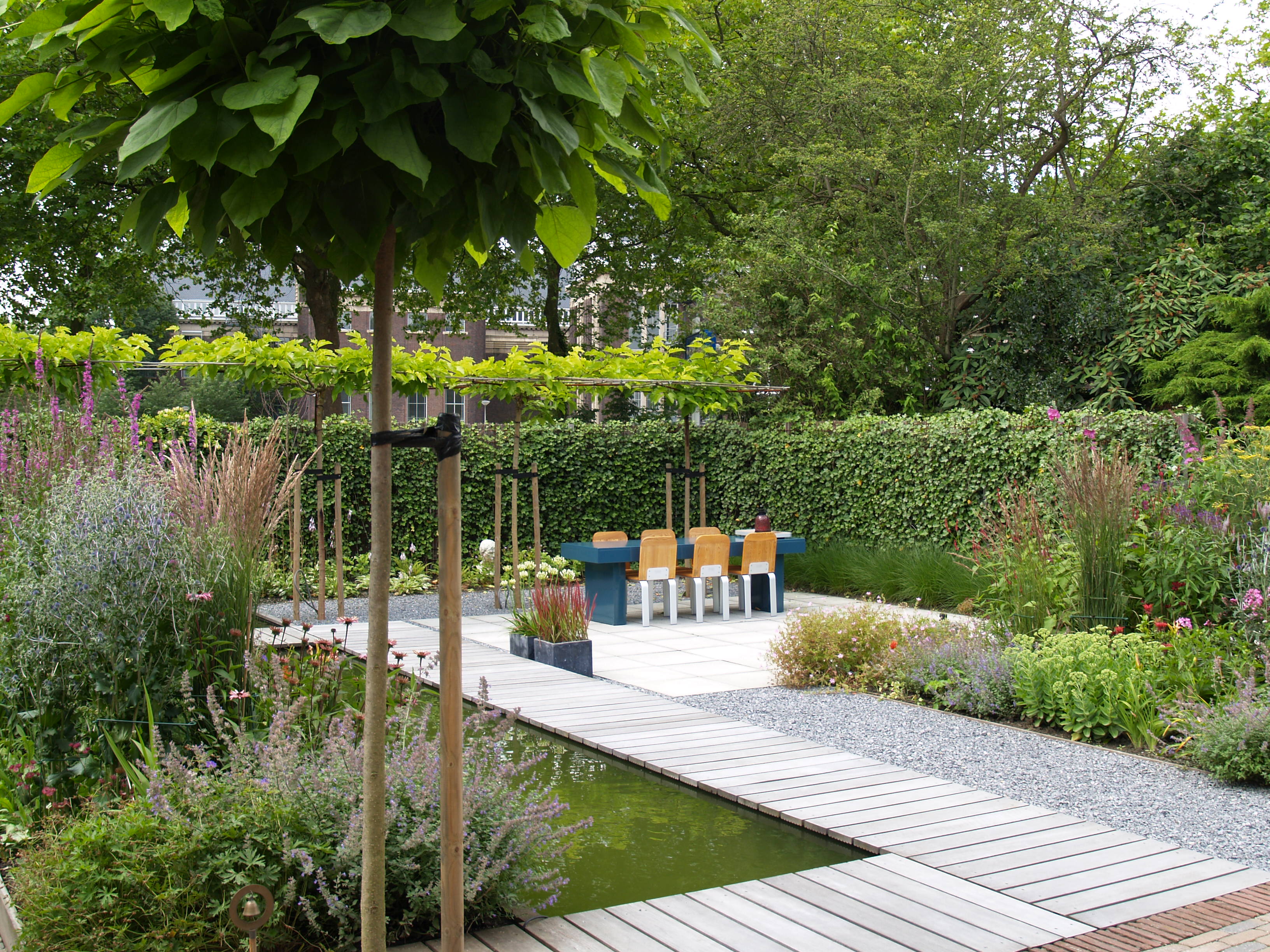 Vlonderpad over vijver naar eethoek met blauwe tafel en oranje stoelen onder Catalpa boom